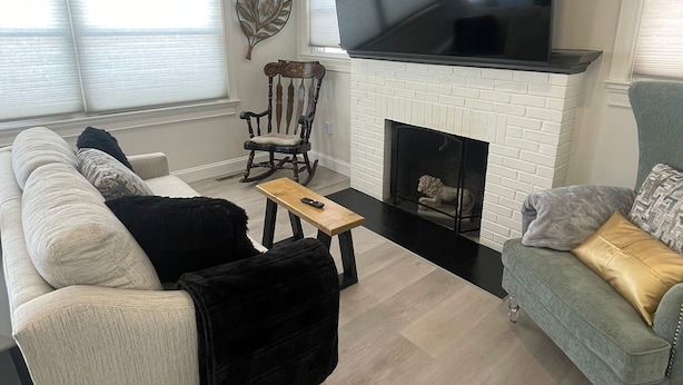 A modern living room with a white brick fireplace, a large flat-screen TV mounted above it, a white sofa, a green armchair, and a wooden coffee table. - Charming 2-bedroom house with WiFi, AC in enjoyable Lancaster (Lancaster)