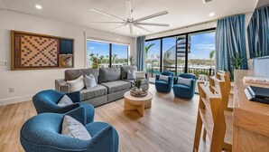 A modern living room with a grey sofa, blue armchairs, a wooden coffee table, and a large window offering a view of a building and palm trees. - New - Private Beach Access/Zip Line & Waterslide (Sarasota)