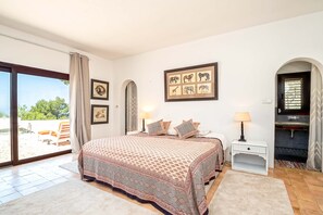 A bedroom with a large bed, a desk, and a chair. There is a window with a view of trees and a pool. - CASA AMAPOLA (Sant Josep de Sa Talaia / San Jose)