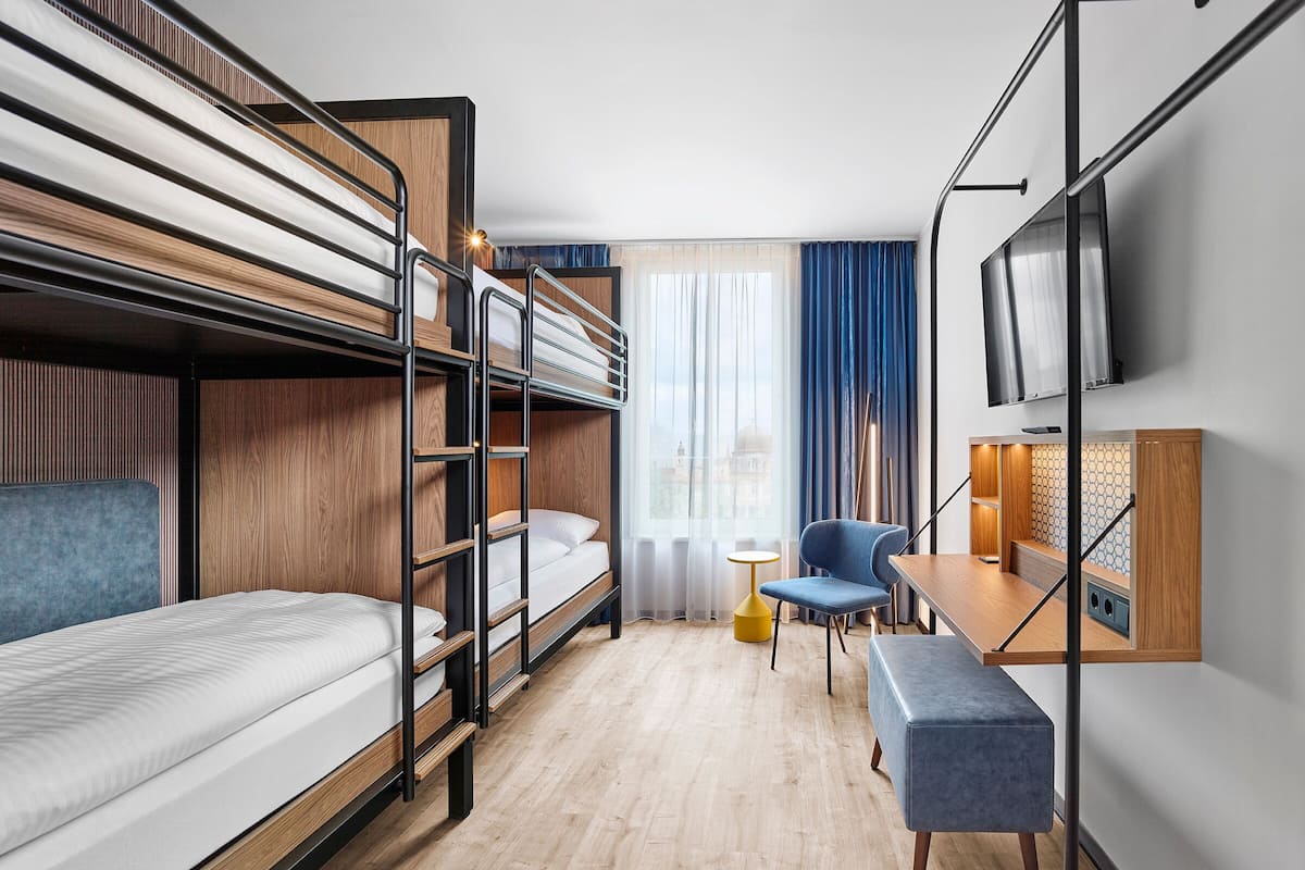 Quarto de hotel com beliches, uma mesa, uma cadeira, televisão e janela com cortinas.