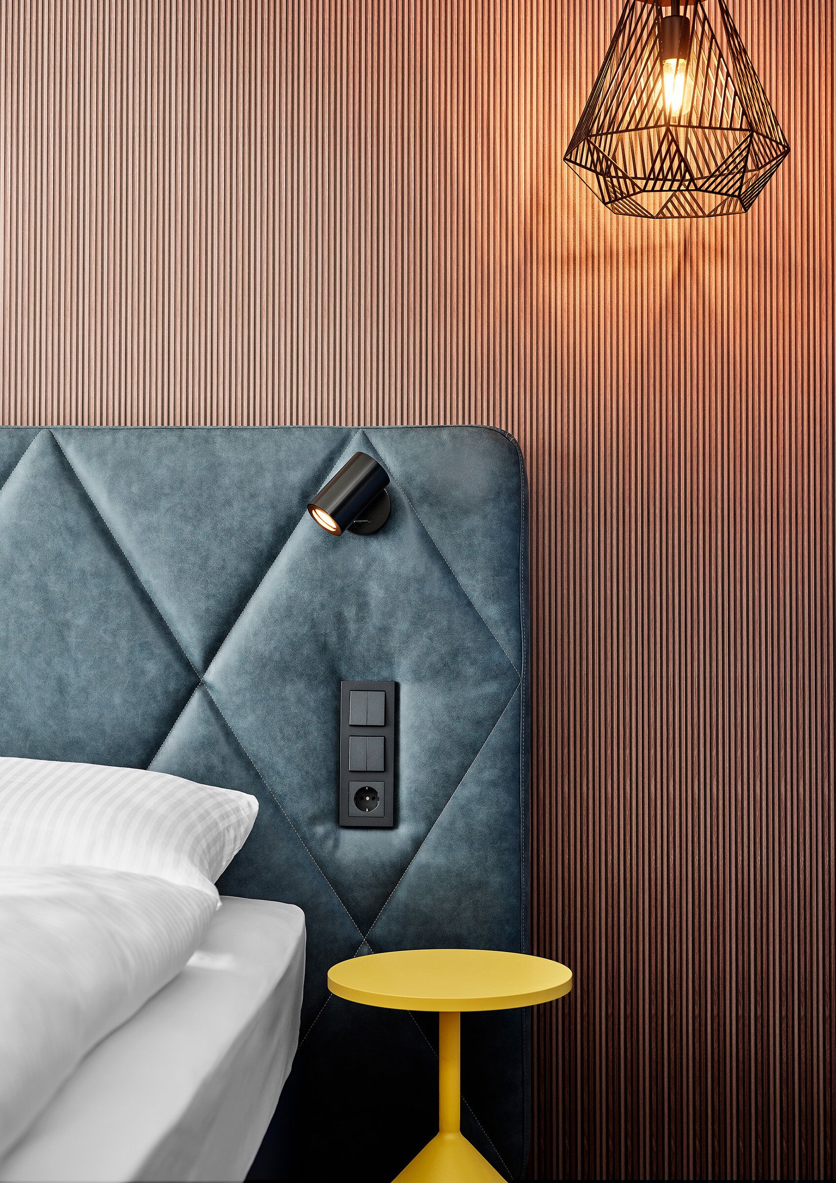 A modern hotel room with a tufted headboard, a bedside table with a lamp, and a small round table.