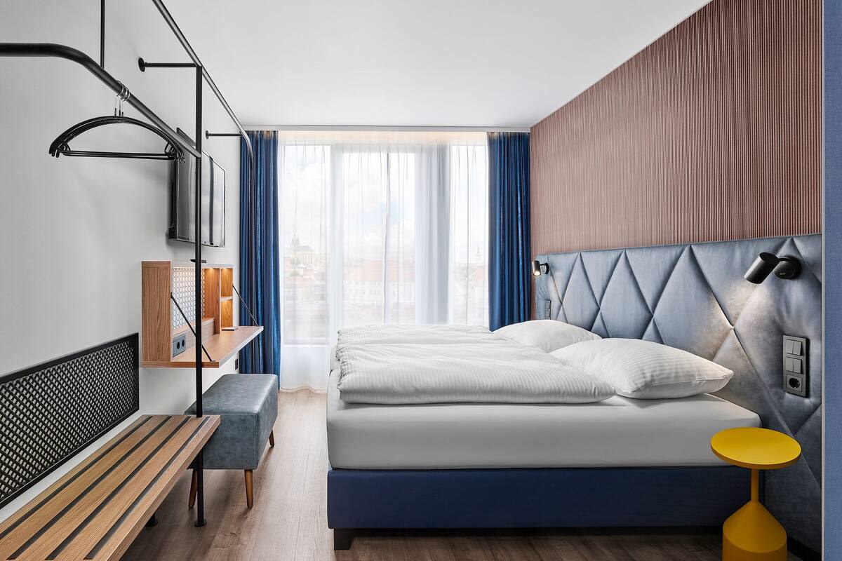 Quarto de hotel moderno com cama, banco, mesa de cabeceira e vista da cidade através de grandes janelas.