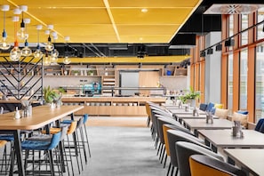 Un café moderne avec des tables et des chaises en bois, un espace cuisine et de grandes fenêtres.