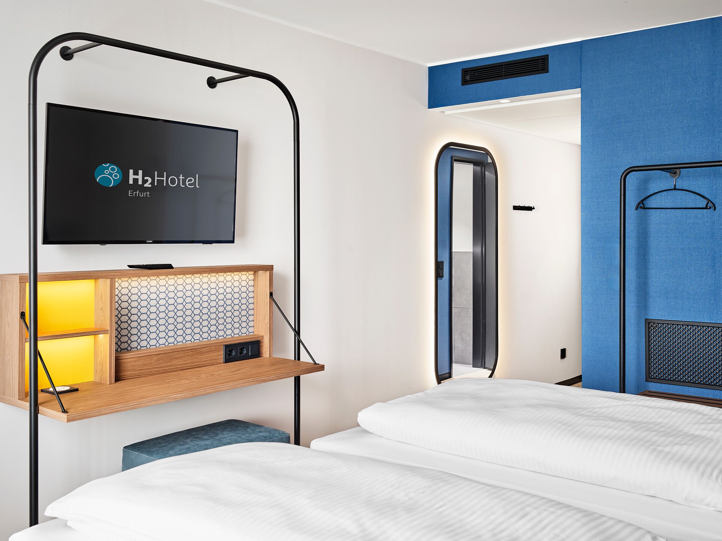 A hotel room with a bed, a TV mounted on a wall, a wooden shelf with a radio, and a mirror.