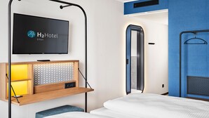 A hotel room with a bed, a TV mounted on a wall, a wooden shelf with a radio, and a mirror.
