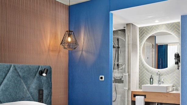 A modern hotel room with a bed, a bathroom with a shower, and a vanity with a mirror.