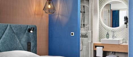 A modern hotel room with a bed, a bathroom with a shower, and a vanity with a mirror.