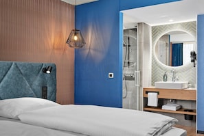 Une chambre d’hôtel moderne équipée d’un lit, d’une table de chevet, d’une lampe fixée au mur, d’un espace douche et d’une coiffeuse avec un miroir.