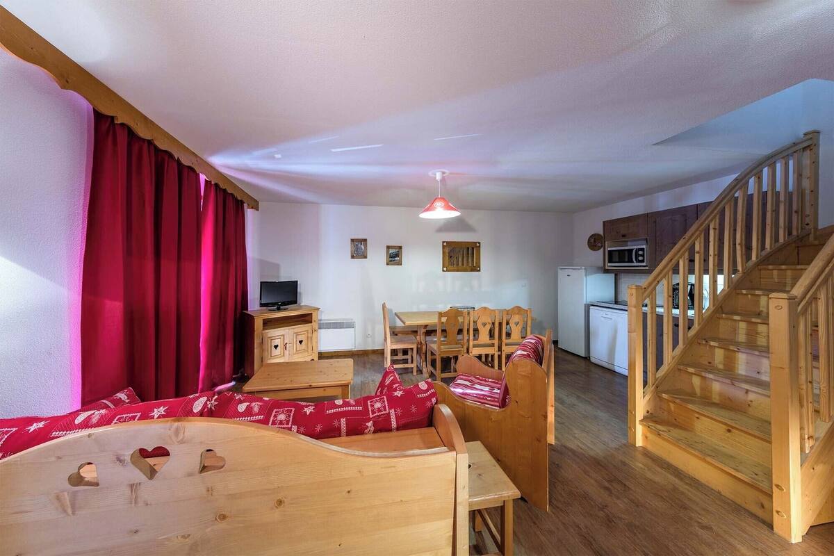 A cozy living room with a wooden sofa, a dining table, and chairs. There's a kitchen area with a microwave and a refrigerator. A wooden staircase leads to another level.