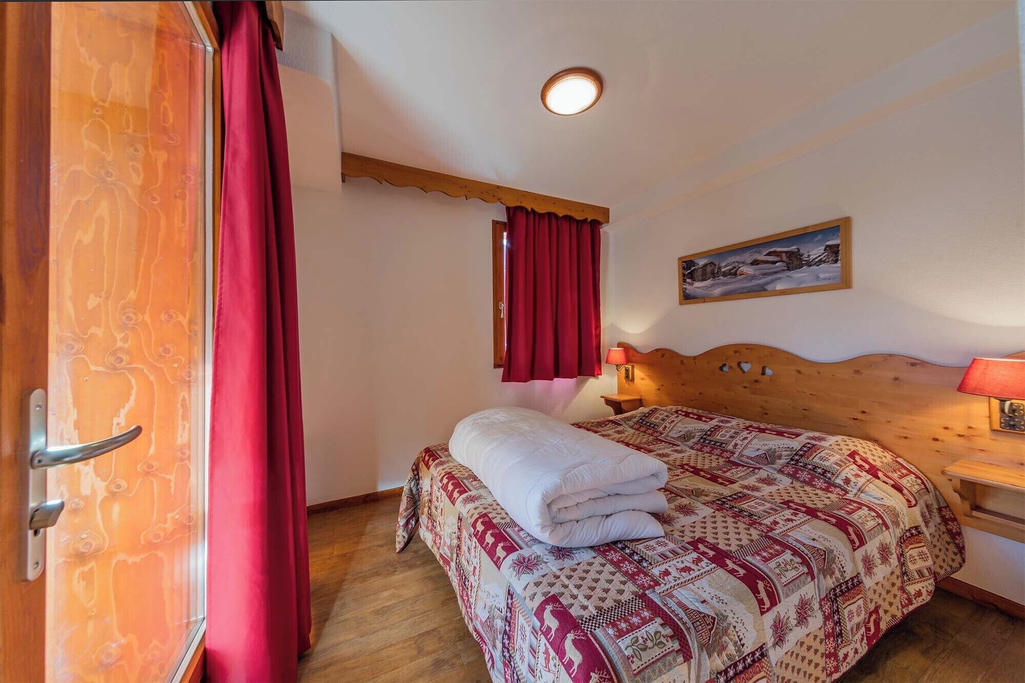 A bedroom with a wooden headboard, a patterned bedspread, a red curtain, and a painting on the wall.