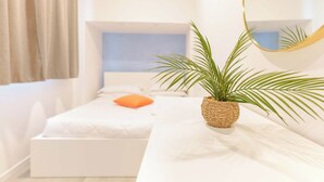 Ein modernes Schlafzimmer mit einem weißen Bett, einem kleinen orangefarbenen Kissen, einem gewebten Blumentopf und einem runden Spiegel auf einer weißen Kommode.