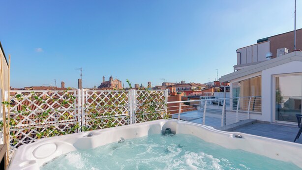 Eine Dachterrasse mit Whirlpool, weißem Gitterzaun und Blick auf Gebäude im Hintergrund.