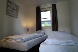 Two single beds with white linens, a large window, and a wall-mounted light fixture.