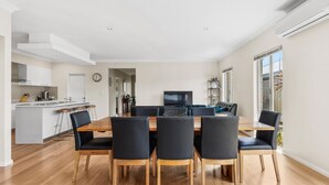 A modern dining area with a wooden table and black chairs, a kitchen in the background, and large windows providing natural light.