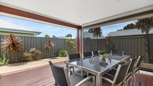 A modern outdoor patio with a glass table and chairs, surrounded by a wooden deck and a view of a backyard with a fence and plants.