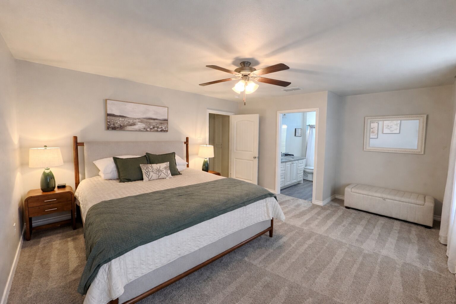 Une chambre à coucher comprenant un lit, des tables de chevet, un ventilateur de plafond, un banc et une salle de bain visible à travers une porte ouverte.
