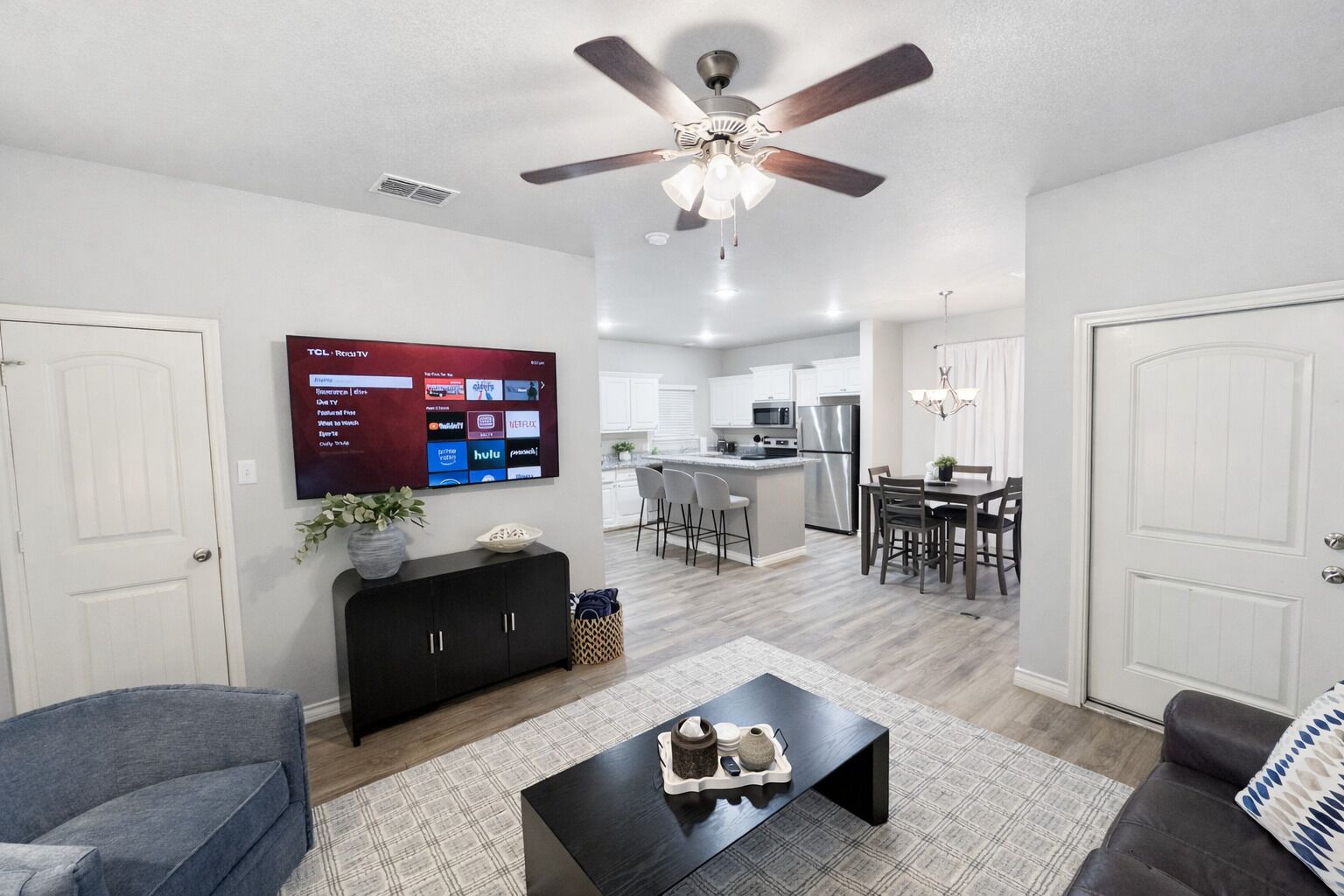 A modern living room with a flat-screen TV, a ceiling fan, a dining area, and a kitchen in the background.
