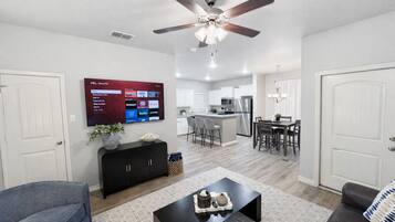A modern living room with a flat-screen TV, a ceiling fan, a dining area, and a kitchen in the background.