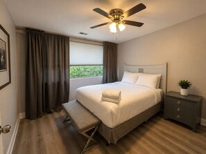 A bedroom with a bed, bedside table, bench, ceiling fan, window with curtains, and a framed picture on the wall.