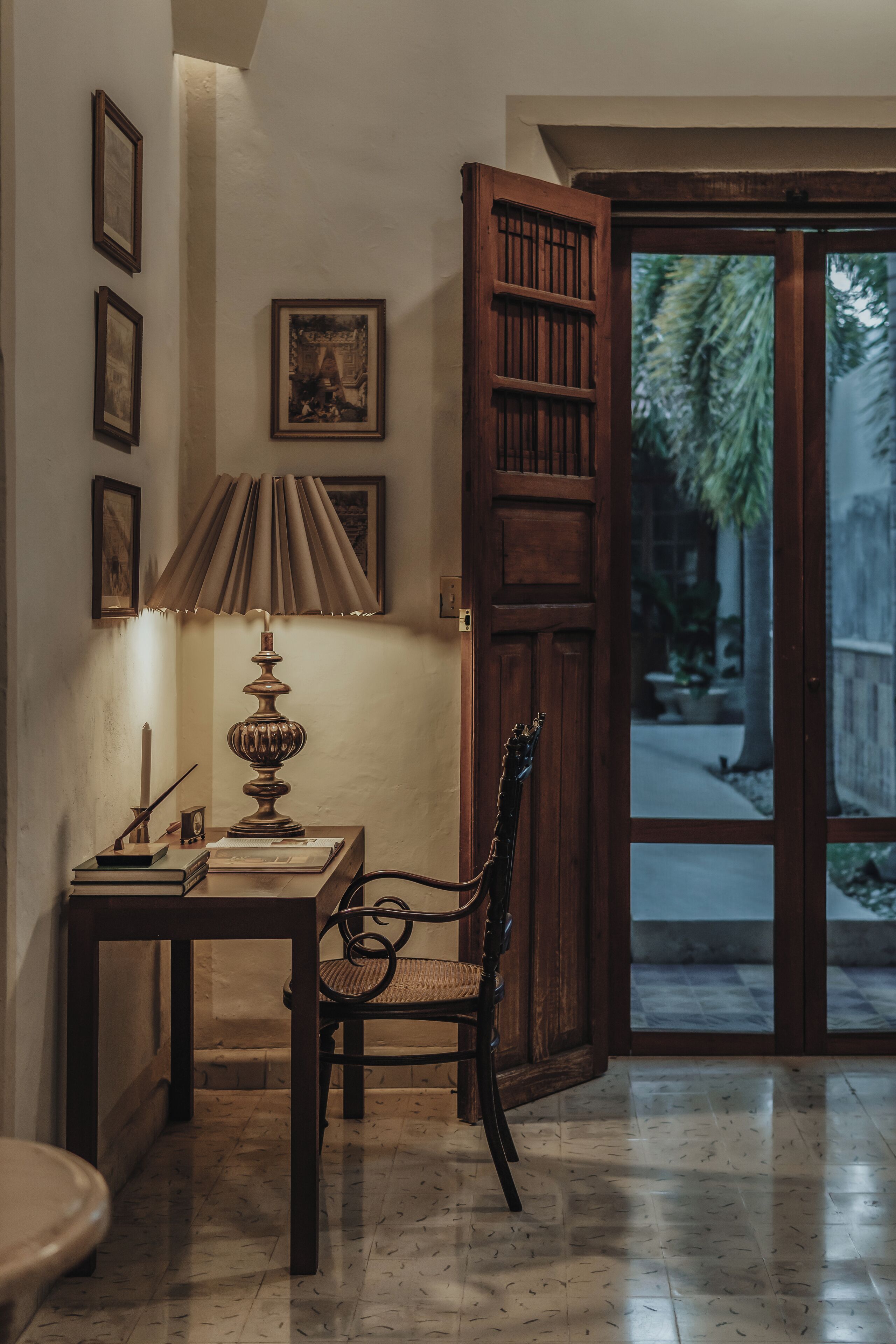 A room with a wooden door, a table with a lamp, a chair, and framed pictures on the wall.