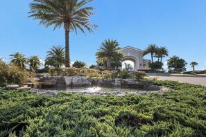 A landscaped garden with a water feature, palm trees, and a building with an arched entrance. - Enjoy Resort living in the Upscale Lakewood National Golf Country Club community (Bradenton)