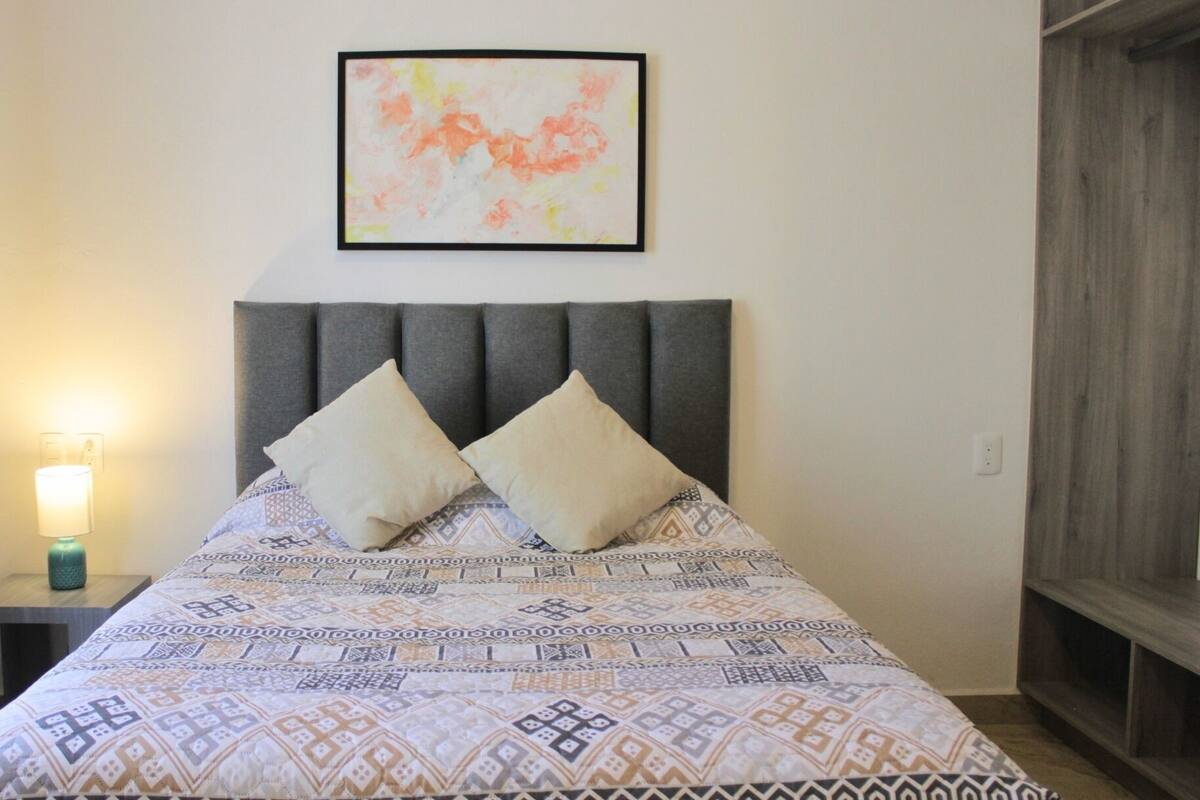 Un dormitorio con una colcha estampada, dos almohadas, un cabecero, una lámpara de noche y un cuadro en la pared.