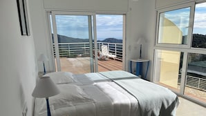 Un dormitorio con cama, mesita de noche, lámpara y vistas al exterior a través de amplias ventanas.