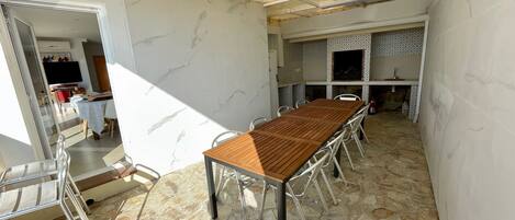 A spacious room with a long wooden table and metal chairs, a stone floor, and a tiled backsplash in the kitchen area.