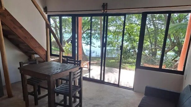 Une pièce avec une table et des chaises en bois, un canapé de couleur sombre et de grandes portes-fenêtres coulissantes donnant sur un espace extérieur verdoyant avec vue sur le ciel.