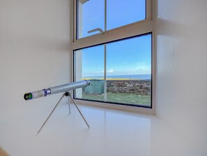 Un telescopio sobre un trípode en una habitación con un amplio ventanal que da a una zona verde y un muro de piedra.