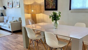Un salon moderne avec un canapé blanc, une table à manger en verre entourée de quatre chaises, un vase rempli de fleurs et un grand tableau abstrait accroché au mur.