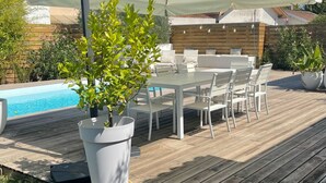 Un patio au bord de la piscine, avec une table et des chaises blanches, un grand parasol et une plante en pot.