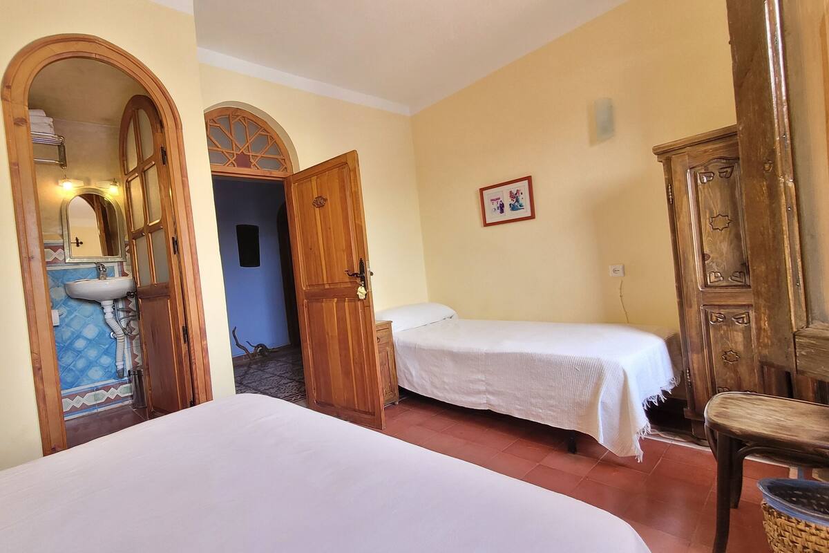 A room with two beds, a wooden wardrobe, a bathroom with a sink and mirror, and a door leading to another room.