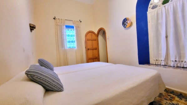 A bedroom with a bed, a window with curtains, a door, and a wall with a decorative plate. - Hotel Gernika (Chefchaouen)