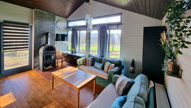 A cozy living room with a stone fireplace, a sofa, a coffee table, and large windows offering a view of the outdoors. - Small country house 2B in the Westerwald. Family-friendly, pet-friendly and close to nature (Welschneudorf)