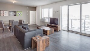 Una sala de estar moderna con un sofá gris, una mesa de centro y un televisor sobre un mueble de madera. Hay una zona de comedor con sillas y una mesa, y ventanas amplias con vistas a la ciudad.