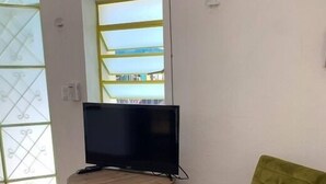 A room with a television on a small table, a green chair, and a view of a building through a window. - Casita in the center of Merida. Only 5 minutes away from La Plancha Park. (Mérida)