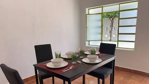 A dining area with a table set for two, chairs, and a window with a view of greenery. - Casita in the center of Merida. Only 5 minutes away from La Plancha Park. (Mérida)