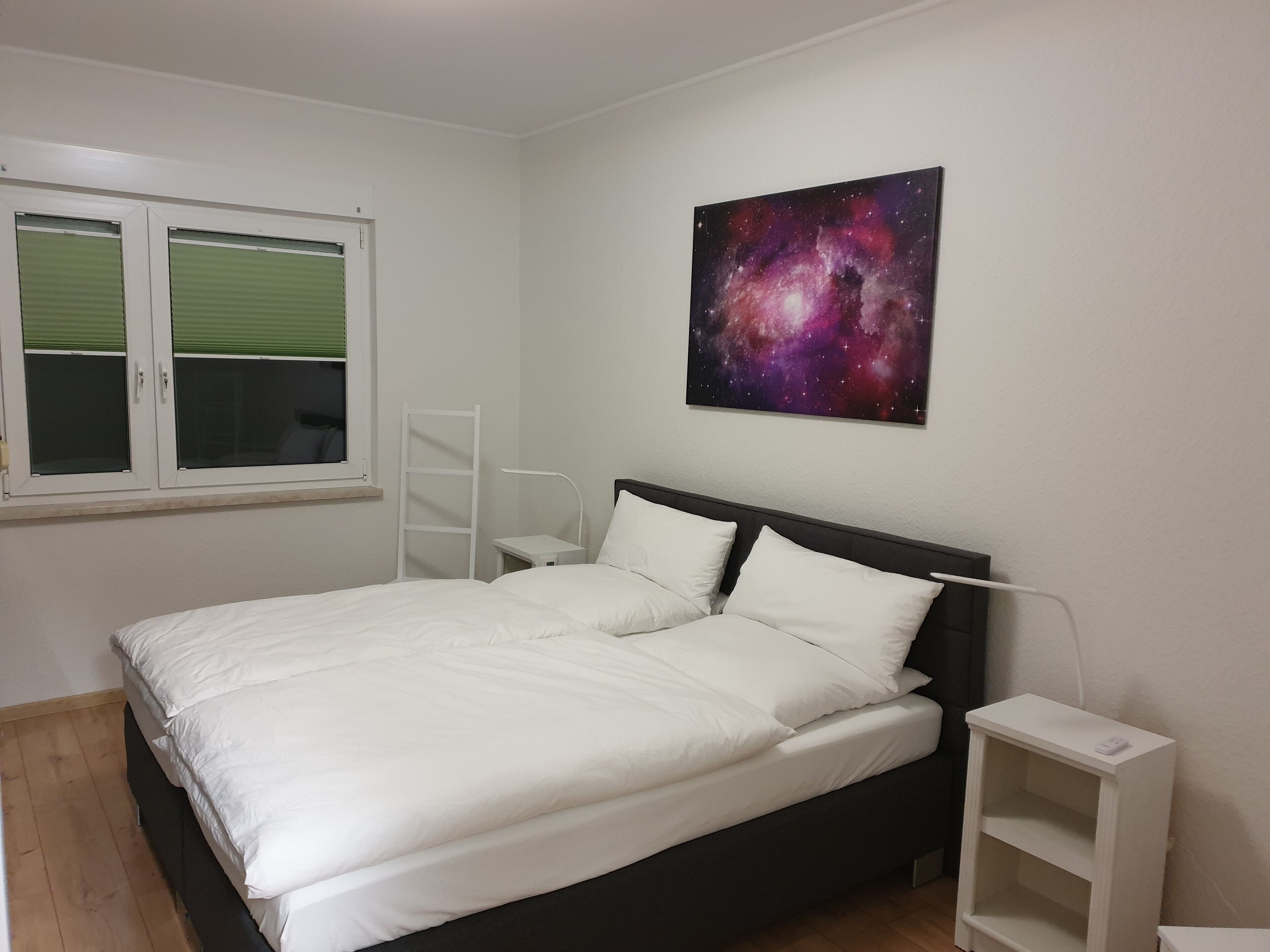 A bedroom with a bed, a nightstand, a wall-mounted lamp, a window with blinds, and a galaxy-themed wall art piece.