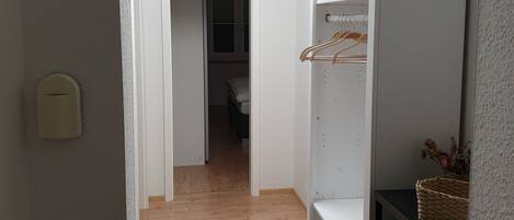 A narrow hallway with wooden flooring, a white wardrobe with shelves and a hanging rod, and a door leading to another room.