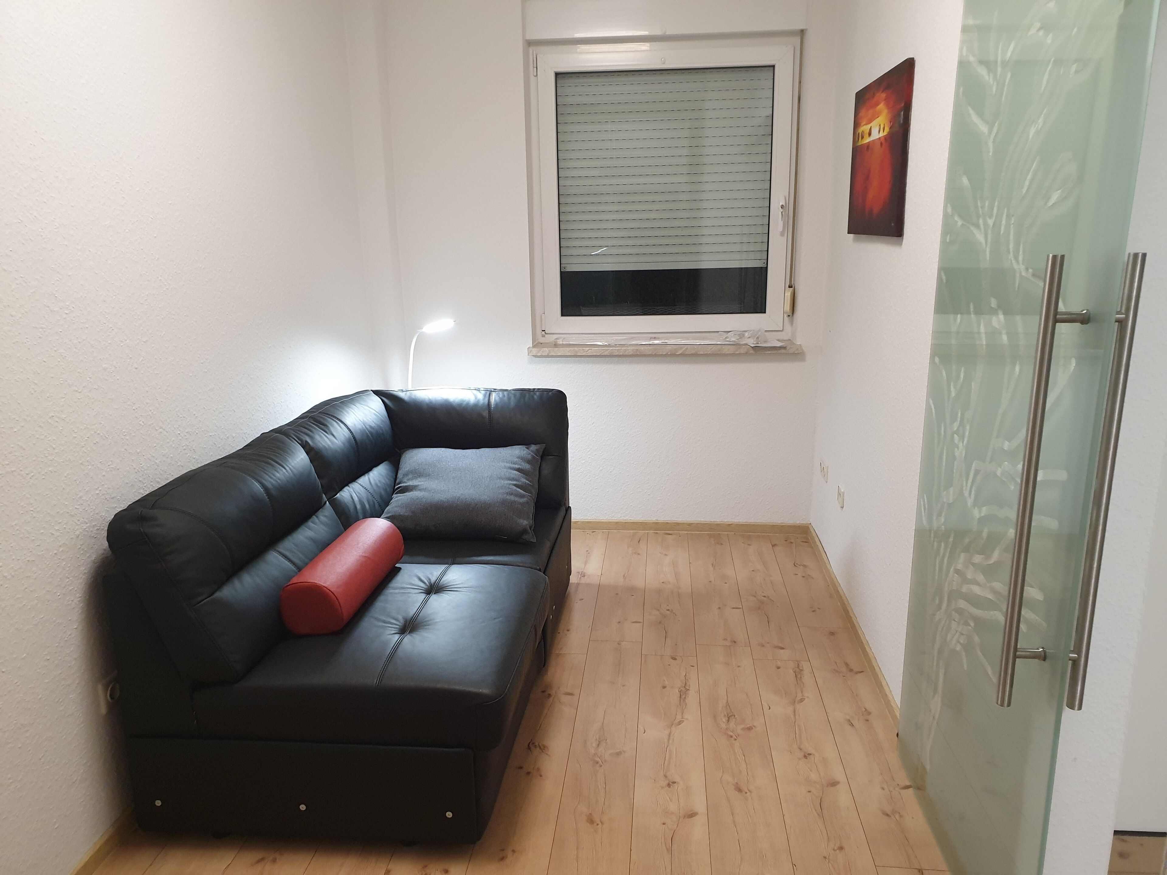 A small, furnished room with a black leather sofa, a red cushion, a wooden floor, a window with blinds, and a glass cabinet.