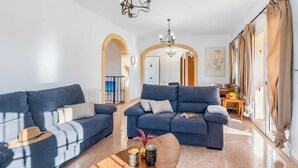 Un salón con dos sofás azules, una mesa de centro con objetos decorativos, una araña y vistas a través de un ventanal corredero.