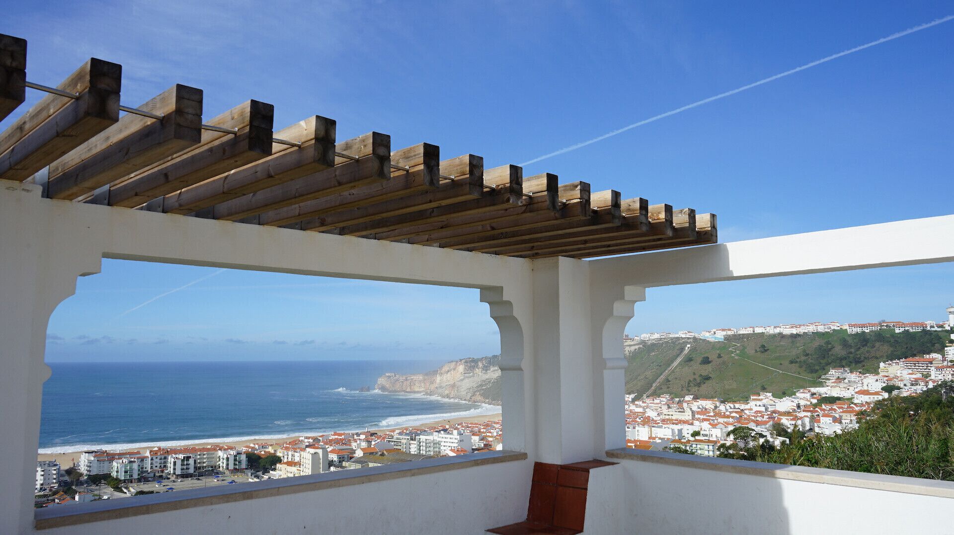 A balcony with a wooden pergola, a view of the sea, and a coastal town in the background.