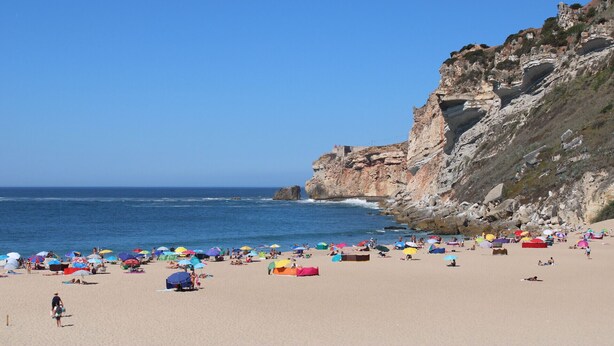 A beach with tents and umbrellas, rocky cliffs, and the ocean. - Lar da Onda - By SCH (Nazaré)