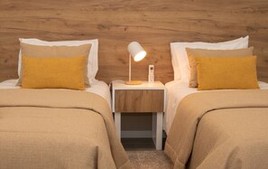 Quarto com duas camas, cada uma acompanhada de uma mesa de cabeceira branca, uma luminária e um controle remoto.