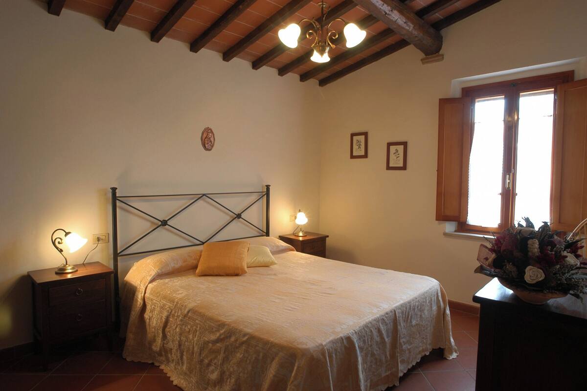 Une chambre à coucher avec un lit, deux tables de chevet avec des lampes, une tête de lit en bois, une fenêtre avec des rideaux et un tableau accroché au mur.
