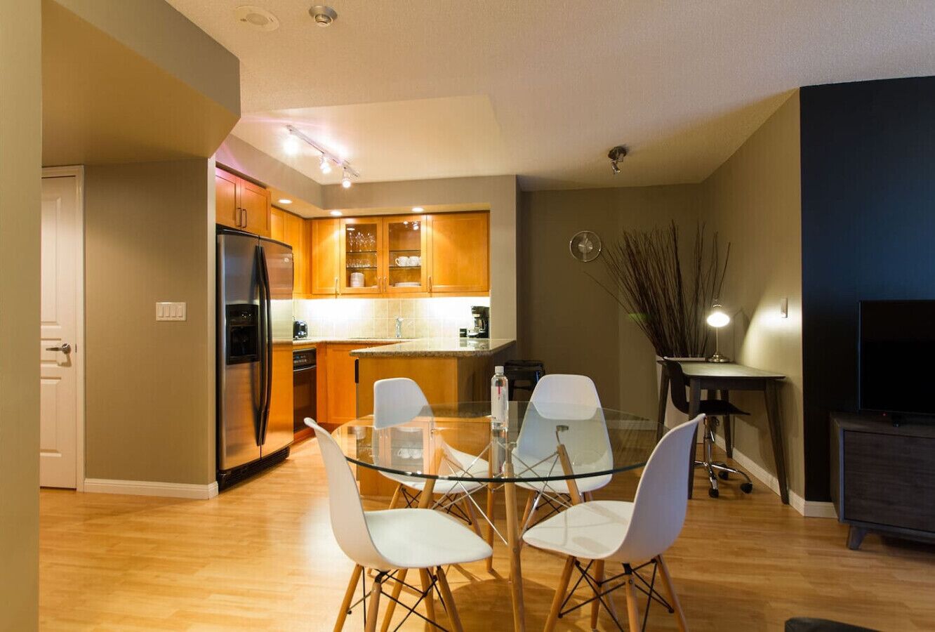 A modern kitchen with wooden cabinets, a stainless steel refrigerator, and a glass dining table with white chairs.