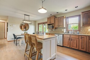 A modern kitchen with wooden cabinets, a central island, a dining area with chairs, and a large window providing natural light.