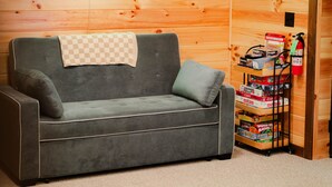 Ein graues Sofa mit einer karierten Decke, ein Neon-Schild mit der Aufschrift 'The Shack' und ein Regal mit verschiedenen Gesellschaftsspielen.