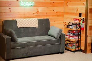 A grey sofa with a checkered throw blanket, a neon sign reading 'The Shack,' and a shelf with various board games.